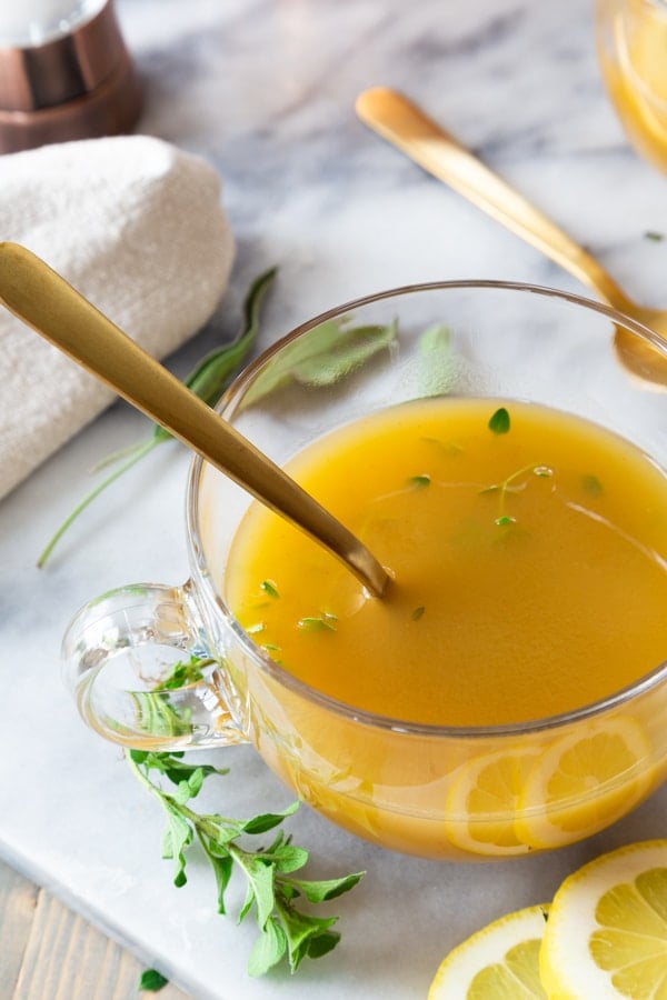 Broth in a clear glass mug with a gold spoon and herbs and lemon slices on a marble board