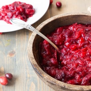 A wood bowl with homemade cranberry sauce and a spoon in the bowl with a white plate with cranberry sauce in the background and loose cranberries on a wood table