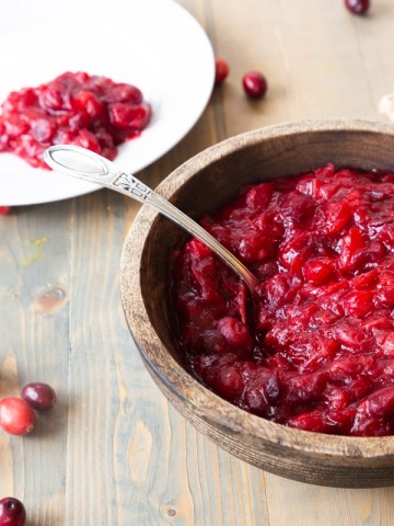 A wood bowl with homemade cranberry sauce and a spoon in the bowl with a white plate with cranberry sauce in the background and loose cranberries on a wood table