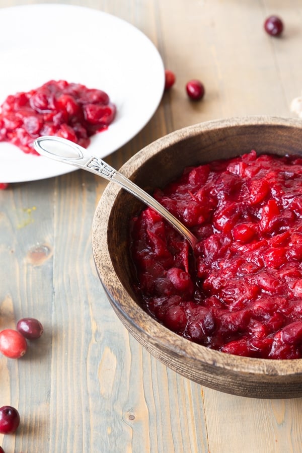 A wood bowl with homemade cranberry sauce and a spoon in the bowl with a white plate with cranberry sauce in the background and loose cranberries on a wood table