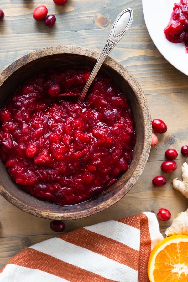 Cranberry sauce in a wood bowl with a spoon in the bowl and an orange and white towel with sliced oranges and cranberries on the wood table