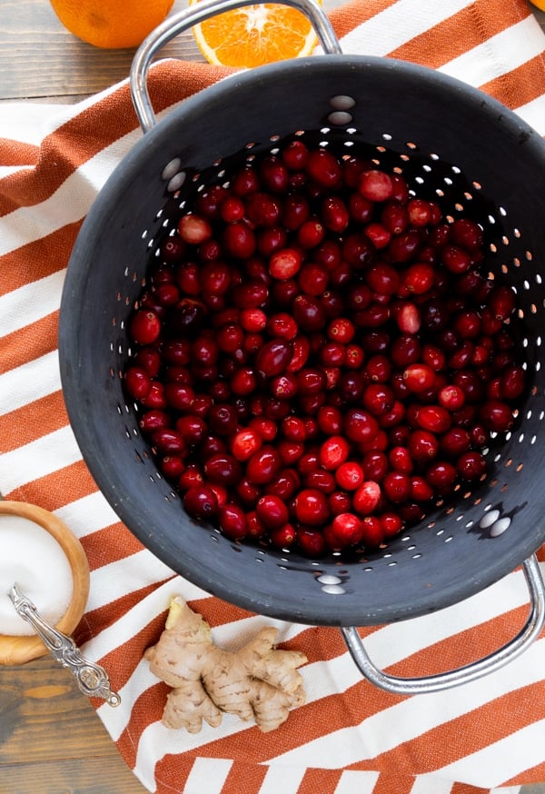 A large gray strainer with fresh cranberries in it on an orange and white striped towel and a wood bowl of sugar and a knob of ginger next to it