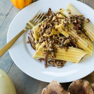 A stack of pumpkin manicotti on a white plate with mushrooms, pecans, and a brown butter sauce on top and a gold fork on the plate with shiitake mushrooms and pumpkins on the side of the plate