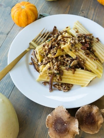 A stack of pumpkin manicotti on a white plate with mushrooms, pecans, and a brown butter sauce on top and a gold fork on the plate with shiitake mushrooms and pumpkins on the side of the plate