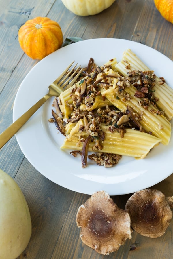 A stack of pumpkin manicotti on a white plate with mushrooms, pecans, and a brown butter sauce on top and a gold fork on the plate with shiitake mushrooms and pumpkins on the side of the plate