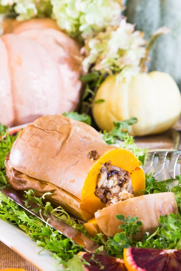 A stuffed butternut squash on a white platter with greens and large pumpkins in the background