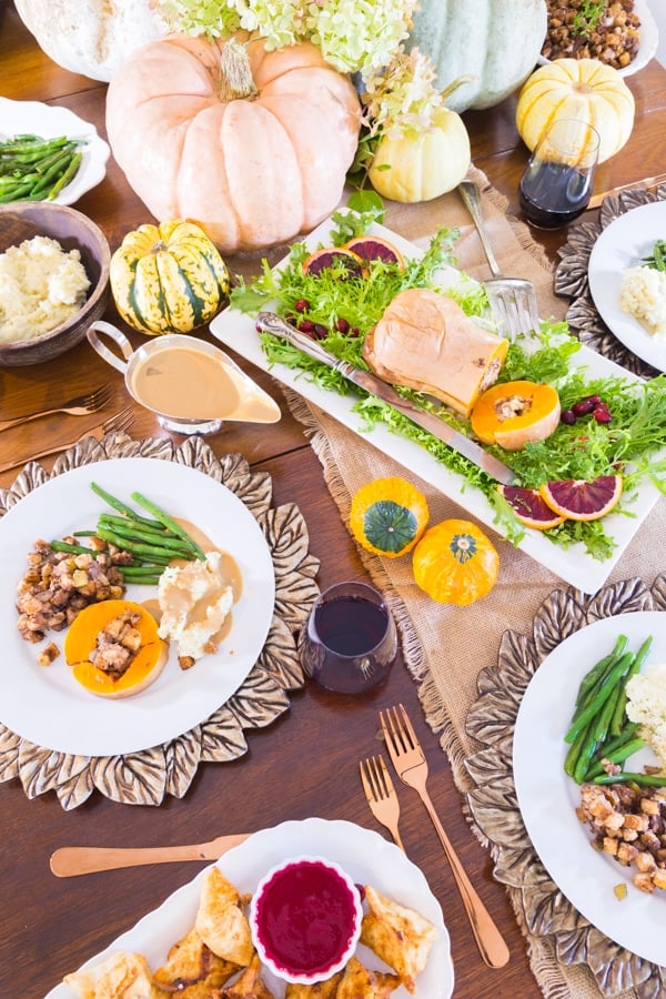 Vegan Thanksgiving table with a white plate with stuffed butternut squash, green beans, mashed potatoes with gravy, stuffing, and a vegan pumpkin centerpiece