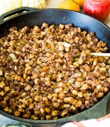 Vegan stuffing in an iron skileet with a spoon in it and a striped towel and apples next to the pan
