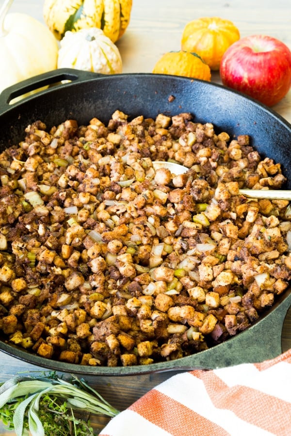 Vegan stuffing in an iron skileet with a spoon in it and a striped towel and apples next to the pan