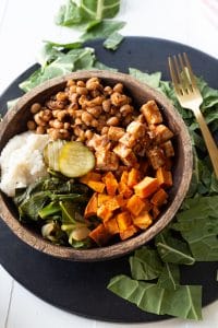 A wood bowl filled with vegan barbecued vegetables on a round black serving platter and greens on the platter created by Veganosity