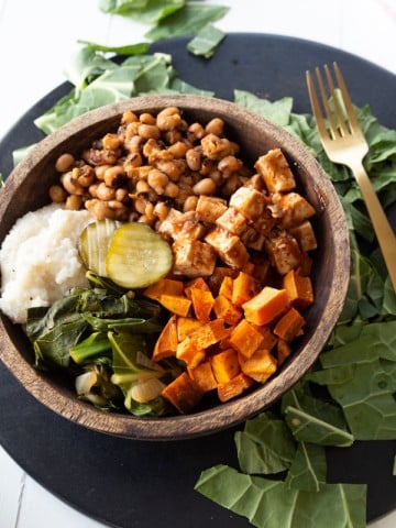 A wood bowl filled with vegan barbecued vegetables on a round black serving platter and greens on the platter created by Veganosity