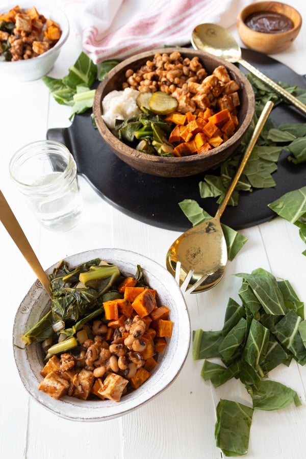 A white bowl of BBQ vegetables with a gold fork and a wood bowl of the BBQ and greens on the table