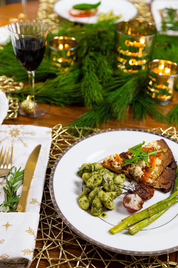 Pesto gnocchi with roasted vegetables and a slice of pizza on a white plate with a silver rim and a glass of red wine and a folded white and gold snowflake napkin with a gold fork and knife and a sprig of rosemary on the side