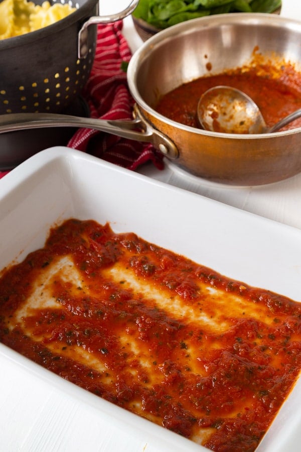 A white ceramic pan with red sauce and lasagna noodles and a copper pot in the background with sauce.