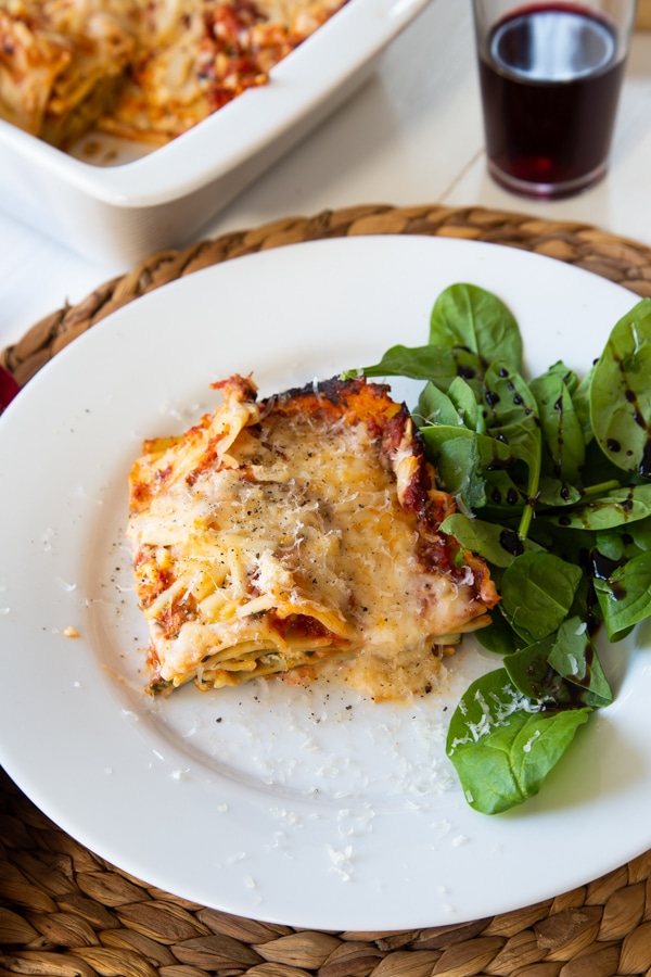 Vegan spinach lasagna and a side salad on a white plate on a braided wicker placemat, and a white ceramic pan of lasagna and a glass of red wine in the background