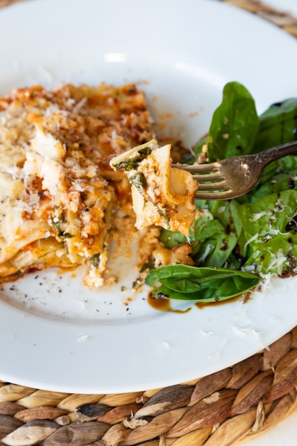 A slice of vegan spinach lasagna on a white plate with a side salad., with a piece of lasagna on a fork