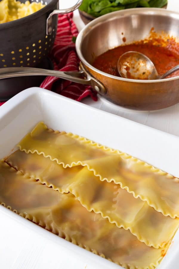 lasagna noodles in a white ceramic pan with a copper pan of sauce in the background.