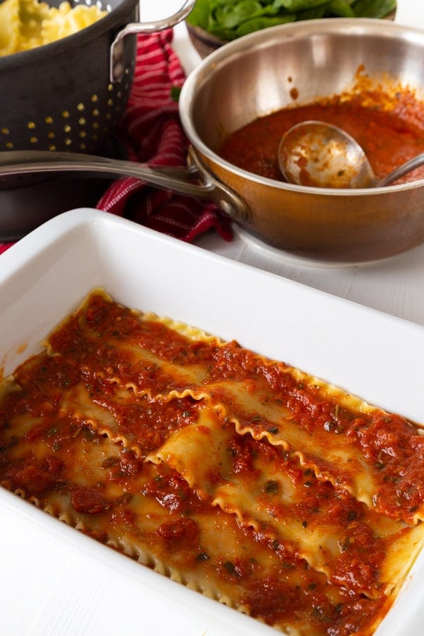 A layer of lasagna noodles and red sauce in a white pan and a copper pan of red sauce. 