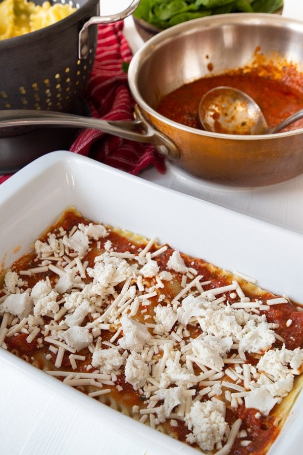 A layer of lasagna noodles, red sauce, and vegan mozzarella cheese in a white pan with a pot of red sauce in the background