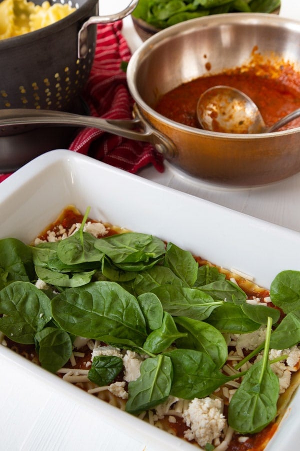 A layer of lasagna noodles, red sauce, vegan mozzarella and ricotta cheese, and baby spinach leaves in a white pan with a pan of sauce in the background.