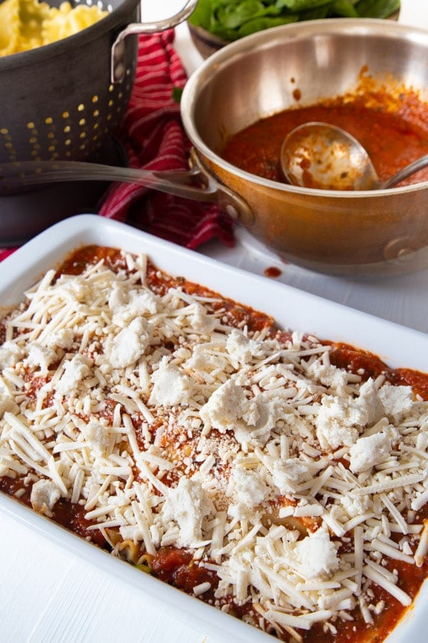 Unbaked vegan lasagna in a white ceramic pan and a copper pot of red sauce in the background.