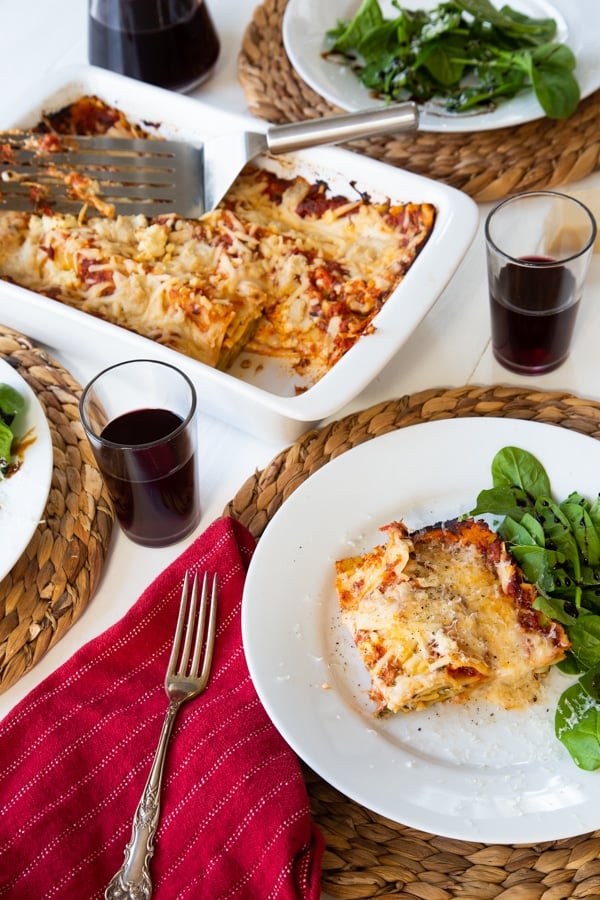 A slice of vegan spinach lasagna on a white plate with a side salad, and another plate of food on a wicker place mat and a white ceramic pan of lasagna in the background