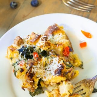 Vegan Strata on a white plate with a gold fork on the edge of the plate and blueberries scattered on a wood table next to the plate