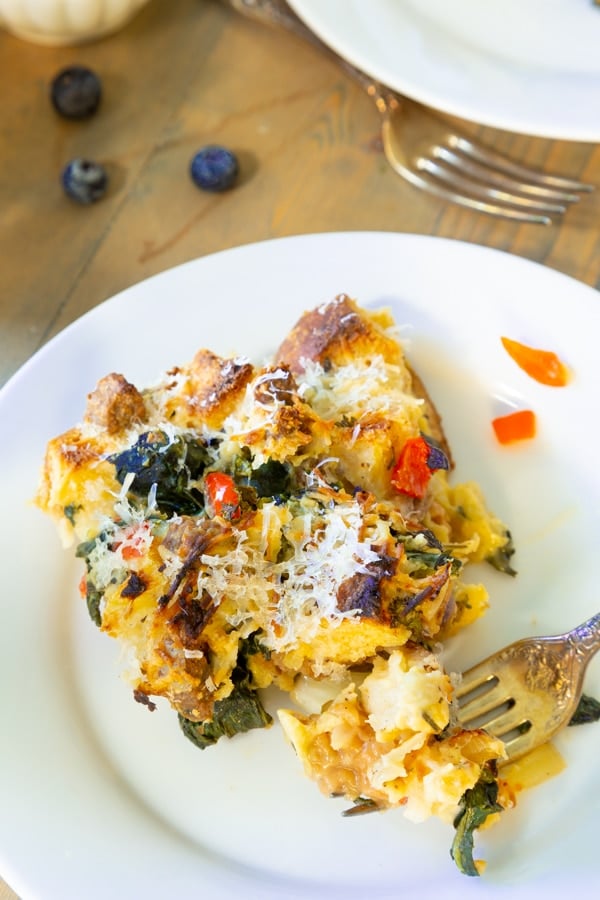 Vegan Strata on a white plate with a gold fork on the edge of the plate and blueberries scattered on a wood table next to the plate