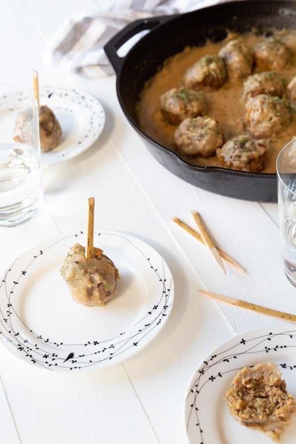 Several small white plates with black birds on the rim and a single Swedish meatball with a toothpick on each plate and a cast-iron skillet filled with vegan Swedish meatballs in the background.