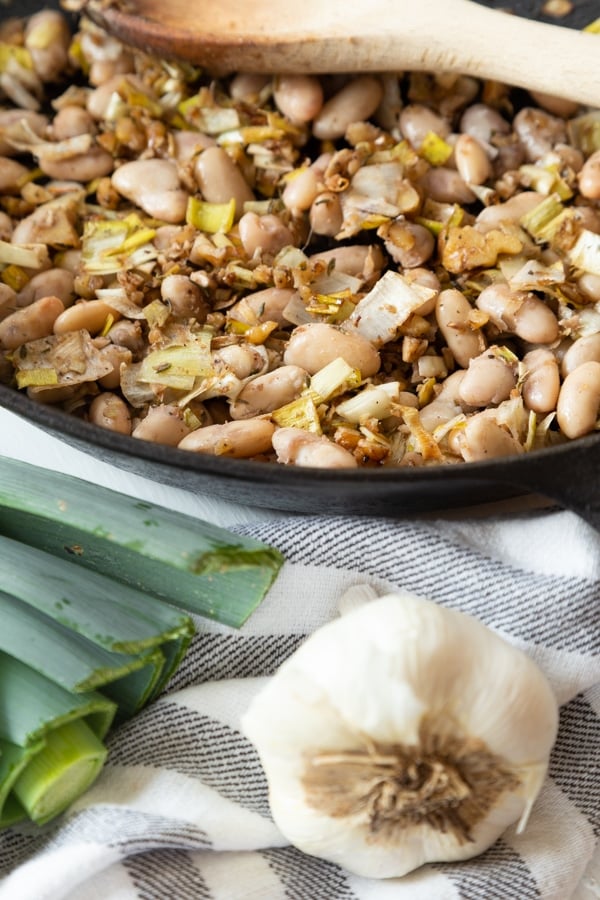 Sauteed leeks, garlic, walnuts, and white beans in a balsamic reduction in an iron skillet with a garlic bulb and leeks next to the pan