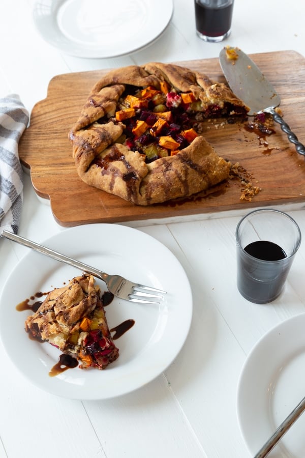 A sweet potato and beet galette on a wood board with a slice out of it on a white plate with a silver fork on the edge and two empty white plates with forks and clear glasses with red wine around the table