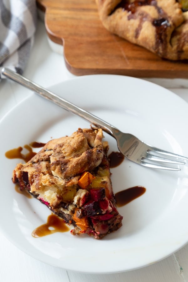 A slice of sweet potato and beet galette on a white plate with a silver fork on the edge of the plate and the whole galette in the backgrountd