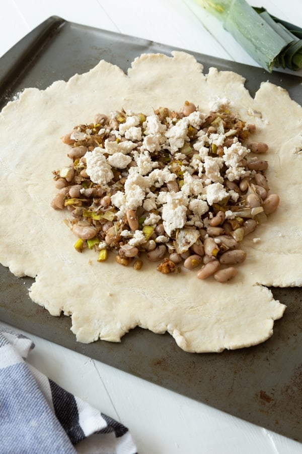 Rolled out pie crust with a bean and leek mixture and vegan ricotta cheese spread on top of the center of the crust on a baking sheet