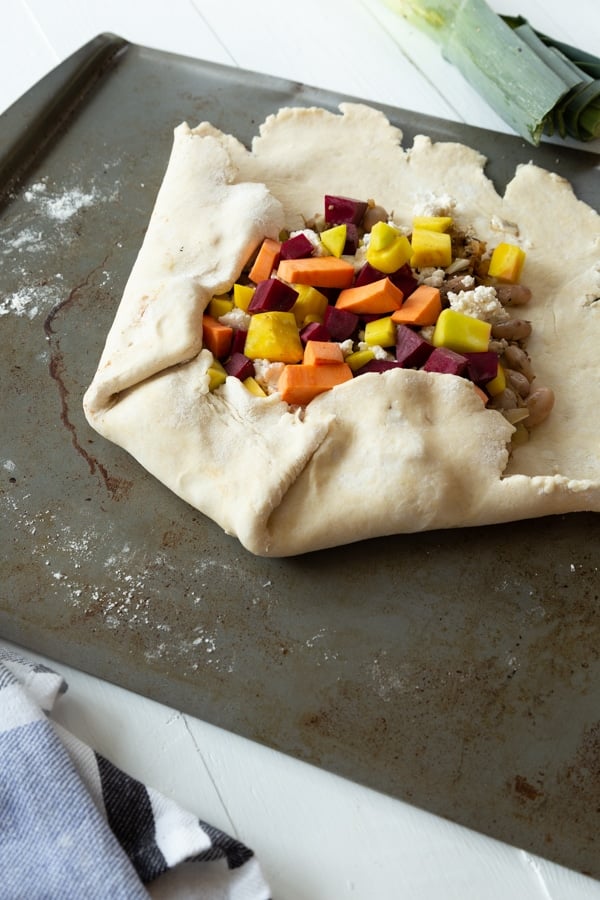 Folding the edges of pie crust over the edges of a sweet potato and beet galette on a baking sheet