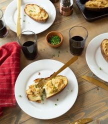 Twice baked ranch potatoes on a wood board with white plates, chives, and glasses of red wine