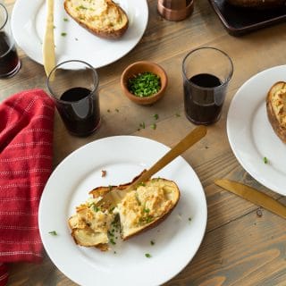 Twice baked ranch potatoes on a wood board with white plates, chives, and glasses of red wine