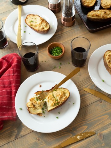 Twice baked ranch potatoes on a wood board with white plates, chives, and glasses of red wine