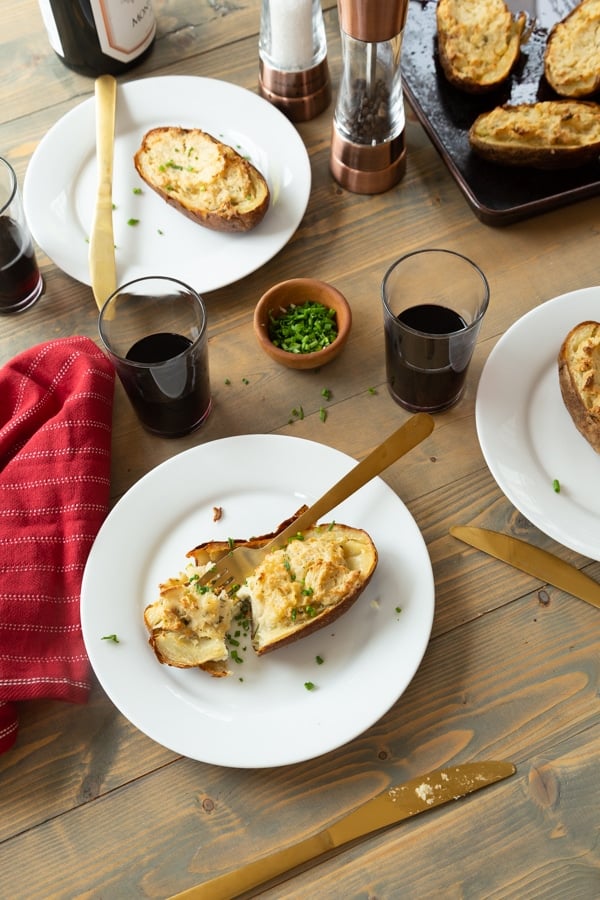 Twice baked ranch potatoes on a wood board with white plates, chives, and glasses of red wine