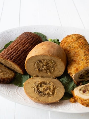 Three vegan turkey roasts on a white platter on greens, sliced on the ends showing the stuffing in the center