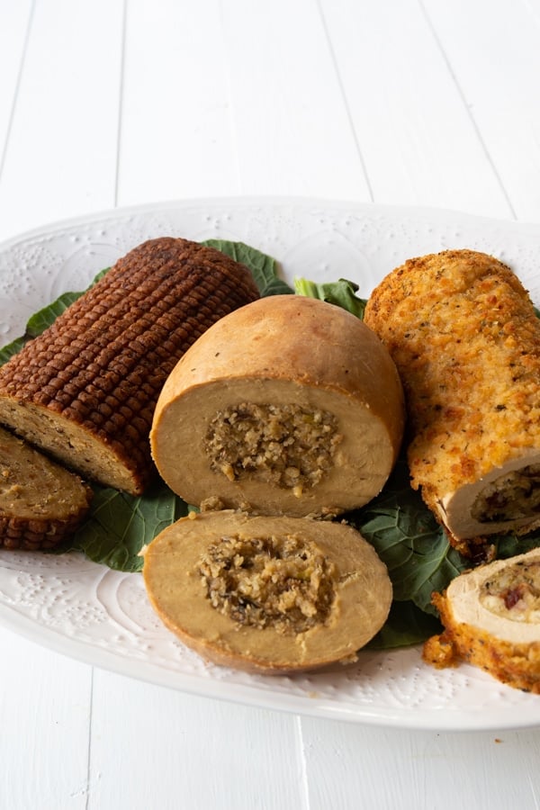 Three vegan turkey roasts on a white platter on greens, sliced on the ends showing the stuffing in the center