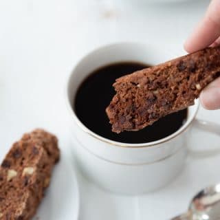 Chocolate chip and pecan biscotti being dipped into a cup of coffee in a white cup with a white plate of biscotti next to it and a silver spoon on the white wood table
