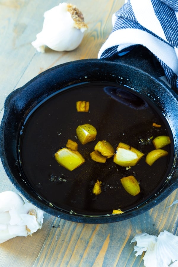 A pan of roasted garlic in olive oil with a blue and white towel wrapped around the handle and garlic cloves next to the pan.