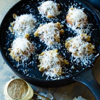 An iron pan with roasted mushrooms covered in Parmesan and a gold measuring spoon full of breadcrumbs and garlic cloves next to it.