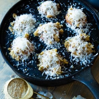 An iron pan with roasted mushrooms covered in Parmesan and a gold measuring spoon full of breadcrumbs and garlic cloves next to it.