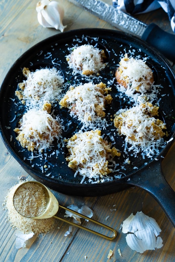 An iron pan with roasted mushrooms covered in Parmesan and a gold measuring spoon full of breadcrumbs and garlic cloves next to it. 