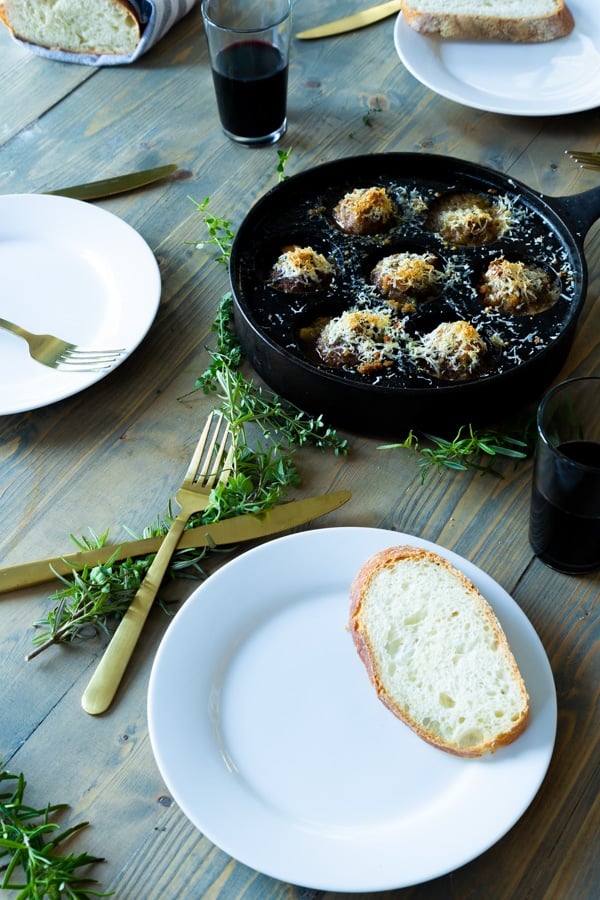 An iron skillet with roasted mushrooms and three white plates with a slice of bread and gold utensils, fresh herbs and glasses of wine around the pan. 