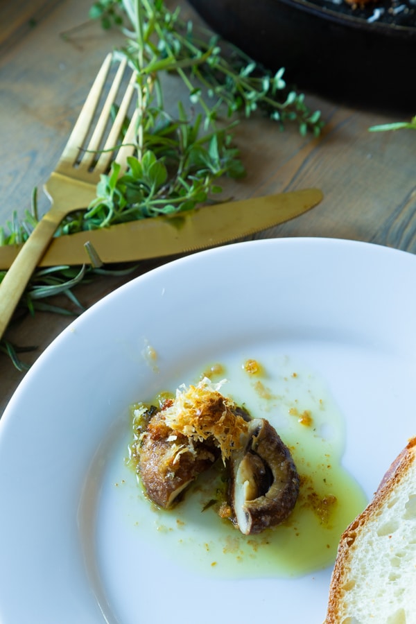 A roasted garlic mushroom on a white plate with a piece of bread and a gold knife and fork and sprigs of fresh thyme next to the plate. 