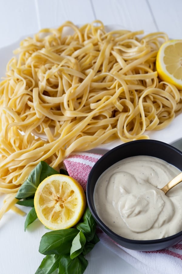 A platter of fettuccine and a bowl of Alfredo sauce with a gold spoon in the black bowl and lemon halves and fresh basil