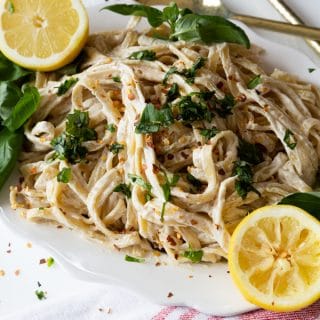 A white platter with vegan Fettuccine Alfredo sprinkled with fresh basil and two lemon halves on the edge of the plate