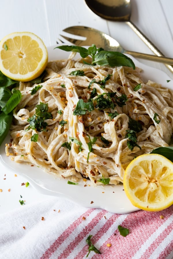 A white platter with vegan Fettuccine Alfredo sprinkled with fresh basil and two lemon halves on the edge of the plate